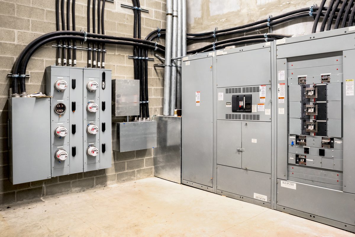 Grey metal switchboard and meter cabinets in a commercial plant room, with cables running up a block wall