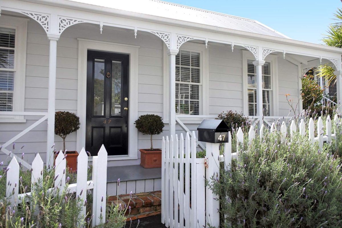 The front of a white weatherboard villa with a fretwork verandah, a black front door and a white picket fence